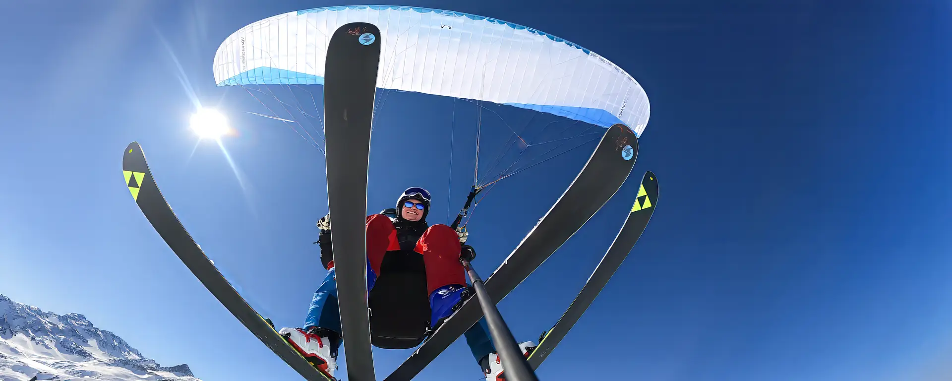 Ski & Fly Tandemflug in Davos von unten: Passagier und Pilot mit Ski an den Füssen in der Luft, darüber der Gleitschirm vor blauem Himmel